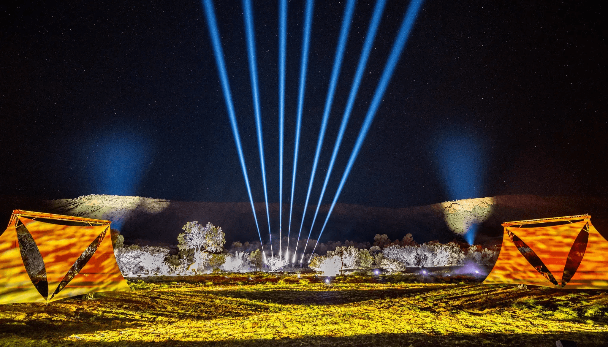Night scene with seven blue searchlight beams rising over illuminated desert, orange-lit fabric shelters on left and right.